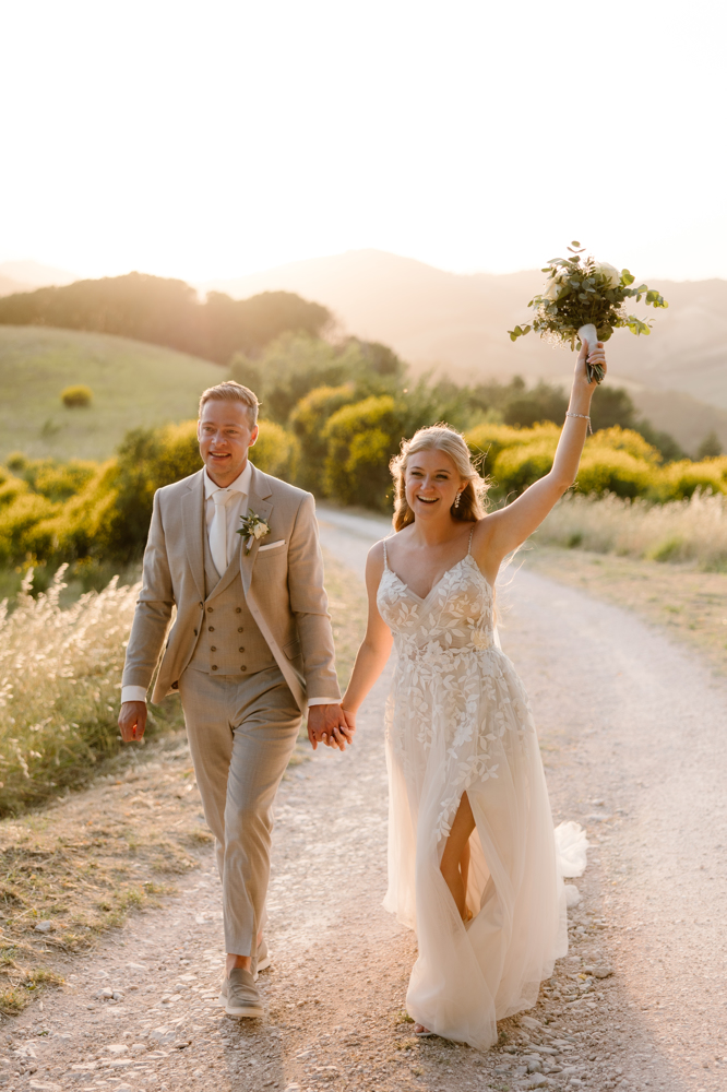 real bride yvon – romantische A-lijn trouwjurk met fijne bloemenapplicaties en elegante split – bruid en bruidegom wandelen hand in hand over een landelijk pad in de avondzon; Yvon straalt terwijl ze haar boeket feestelijk in de lucht houdt